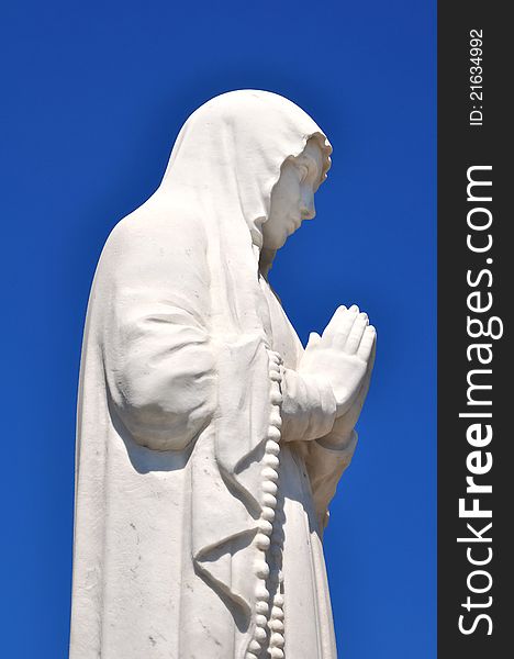 White statue of the Virgin with hands joined in prayer on blue sky background. White statue of the Virgin with hands joined in prayer on blue sky background