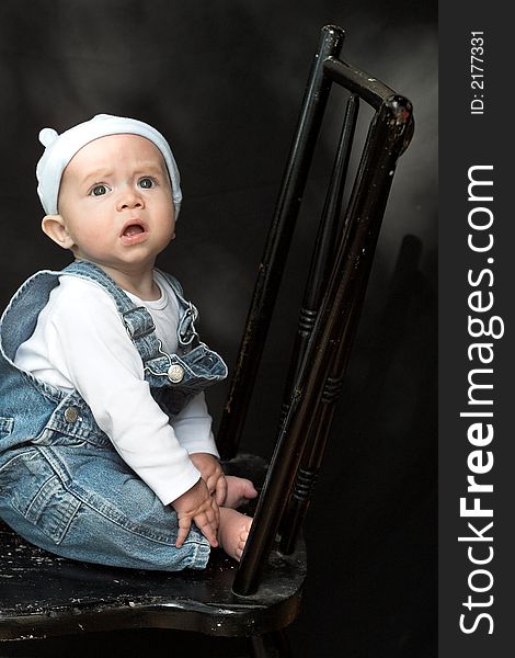 Image of adorable baby wearing denim overalls sitting on a black chair. Image of adorable baby wearing denim overalls sitting on a black chair