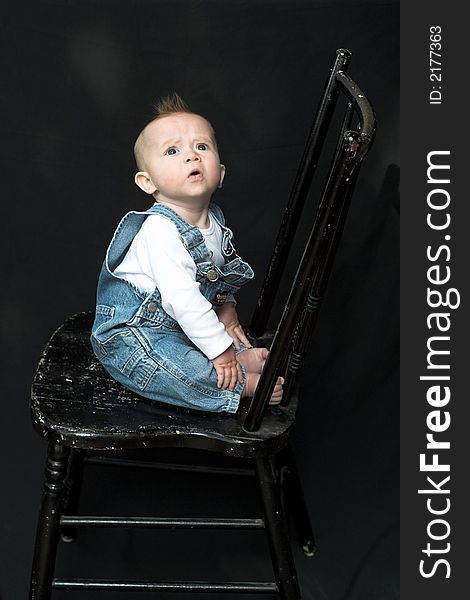 Image of adorable baby wearing denim overalls sitting on a black chair. Image of adorable baby wearing denim overalls sitting on a black chair