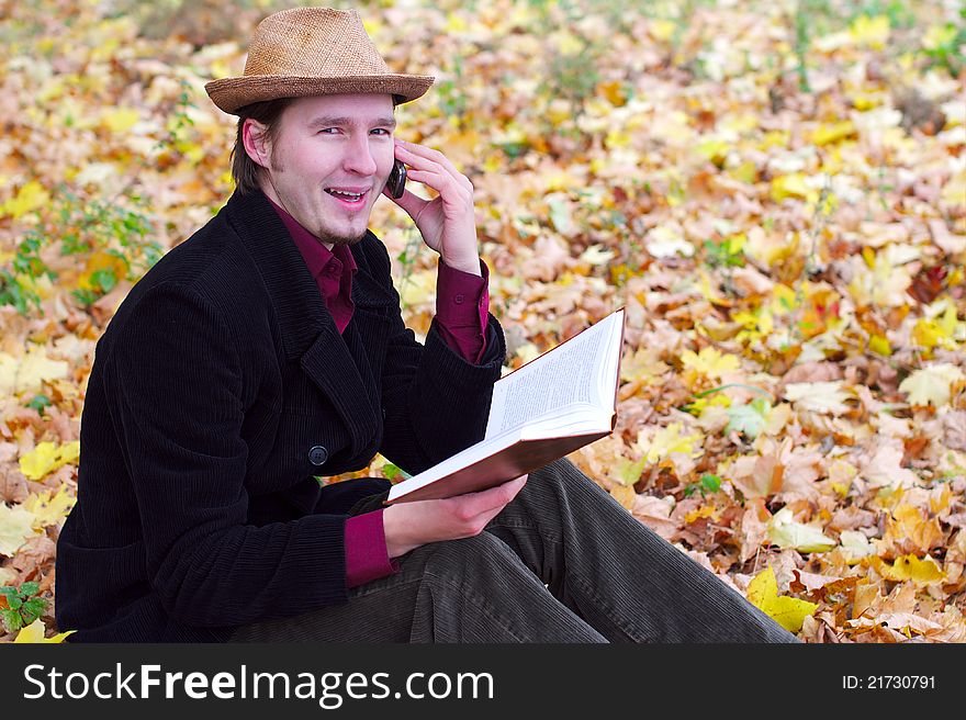 Man with hat is talking a phone and holding a book in autumn leaves background. Man with hat is talking a phone and holding a book in autumn leaves background