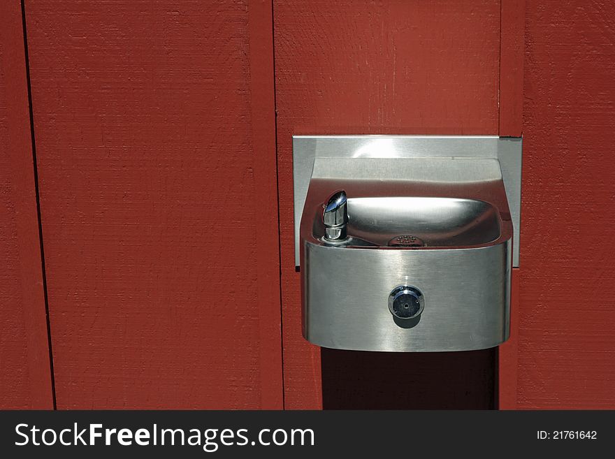 Silver steel water fountain attached to a painted red wood wall outdoors on a sunny day. Silver steel water fountain attached to a painted red wood wall outdoors on a sunny day.