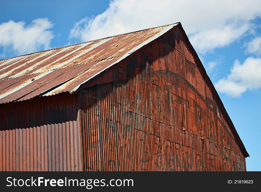 The metal roof and front of an old, rusty, metal building, stands against a blue sky with white clouds. The metal roof and front of an old, rusty, metal building, stands against a blue sky with white clouds.