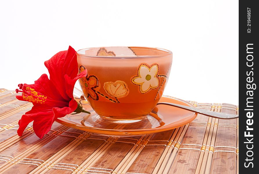 The cup of tea with a beautiful flower of hibiscus on the bamboo mat. The cup of tea with a beautiful flower of hibiscus on the bamboo mat