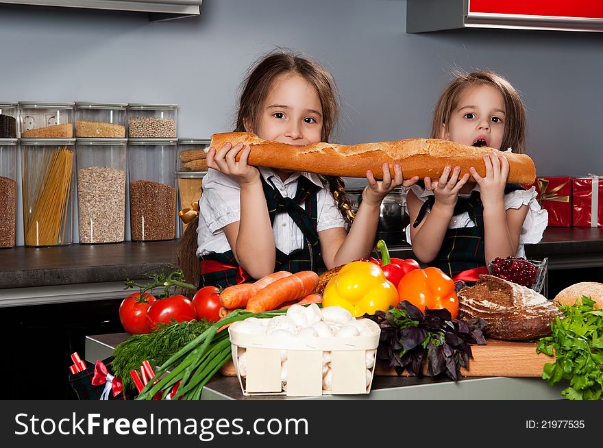 Two little girl in the kitchen of chef uniforms baguette bread bites against the kitchen table with fresh produce