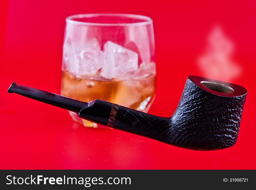 Pipe for smoking tobacco and a glass of whiskey with ice photographed against a red background. Pipe for smoking tobacco and a glass of whiskey with ice photographed against a red background