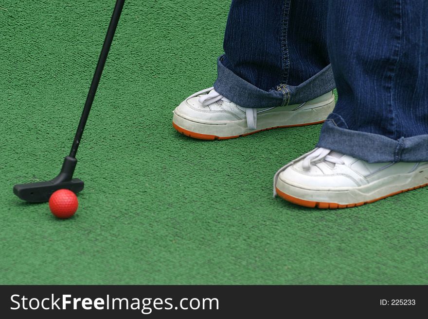 Tennis shoes, putter, and red golf ball at putt putt golf.