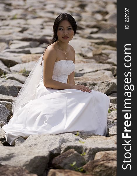 A beautiful asian woman sits on a rocky beach in a wedding dress. A beautiful asian woman sits on a rocky beach in a wedding dress