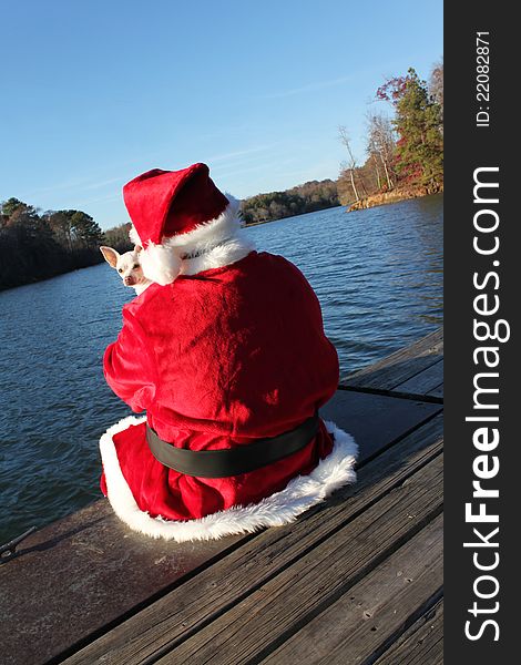 A white chihuahua peers over Santa's sholder as he sits on a dock taking a break from his hectic schedule. A white chihuahua peers over Santa's sholder as he sits on a dock taking a break from his hectic schedule.