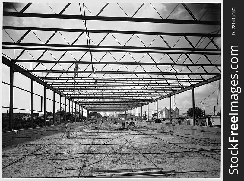 Photograph of a warehouse and office building during construction. The building was constructed by Alco Products, Inc. out of Beaumont using materials provided by John Dollinger Jr., Inc. The steel framing has been completed and workers appear to be sweeping the area, which is a dirt floor. An additional man is walking across a beam at the top of the photograph. Houses and a large building are in the background. Photograph of a warehouse and office building during construction. The building was constructed by Alco Products, Inc. out of Beaumont using materials provided by John Dollinger Jr., Inc. The steel framing has been completed and workers appear to be sweeping the area, which is a dirt floor. An additional man is walking across a beam at the top of the photograph. Houses and a large building are in the background.