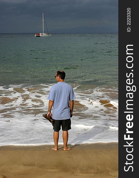 A man stands on the golden sand at the shoreline, gazing at the turquoise blue sea. Perhaps wishing the catamaran offshore is his?. A man stands on the golden sand at the shoreline, gazing at the turquoise blue sea. Perhaps wishing the catamaran offshore is his?