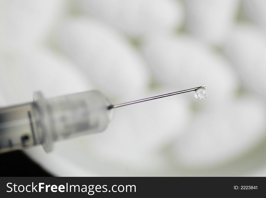 A water drop is at the very end of a syringe with white pills in the background. A water drop is at the very end of a syringe with white pills in the background