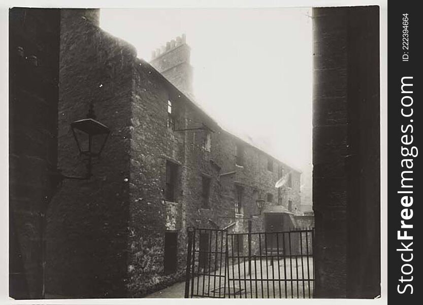 Photograph of a courtyard looking up to a two storey building . There are iron railings and a gate across the left side of the building and iron posts along the  front of the building. There is a street lamp on the left attached to the wall in the foreground. The  courtyard is concrete.

digital.nls.uk/74506932. Photograph of a courtyard looking up to a two storey building . There are iron railings and a gate across the left side of the building and iron posts along the  front of the building. There is a street lamp on the left attached to the wall in the foreground. The  courtyard is concrete.

digital.nls.uk/74506932