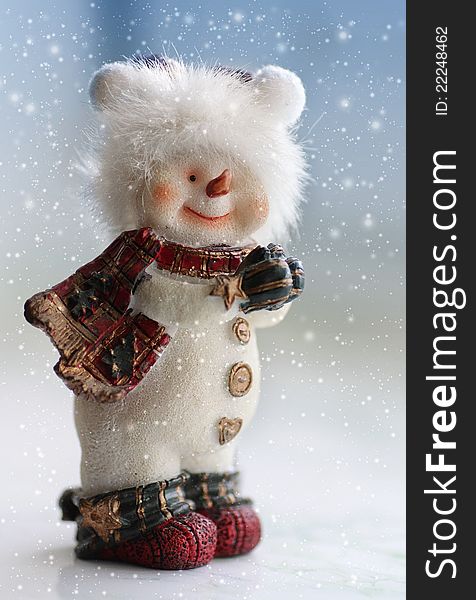 Toy snowman wearing scarf and gloves standing under falling snow on gradient blue background
