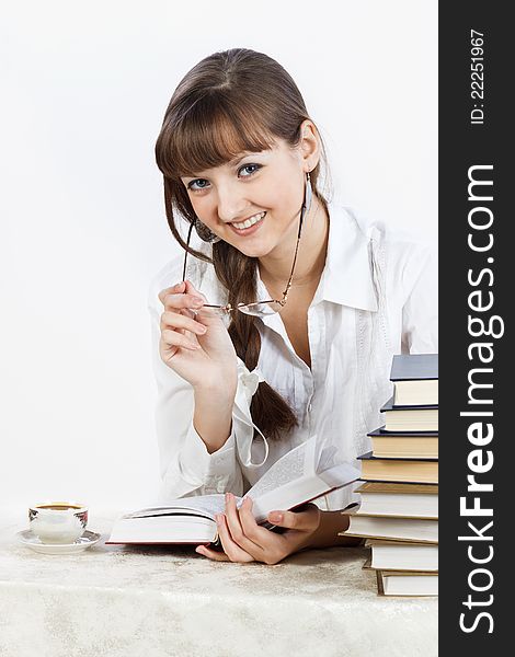 Beautiful smiling girl in a white shirt reading a book while sitting at the table. In her hands she holds the glasses. The girl looks into the camera. By her side there is a cup of coffee. Fragmant of a stack of books can be seen near the edge of photo. Her Hair is braided into a braid. Photographed in a studio against a white background. Beautiful smiling girl in a white shirt reading a book while sitting at the table. In her hands she holds the glasses. The girl looks into the camera. By her side there is a cup of coffee. Fragmant of a stack of books can be seen near the edge of photo. Her Hair is braided into a braid. Photographed in a studio against a white background.