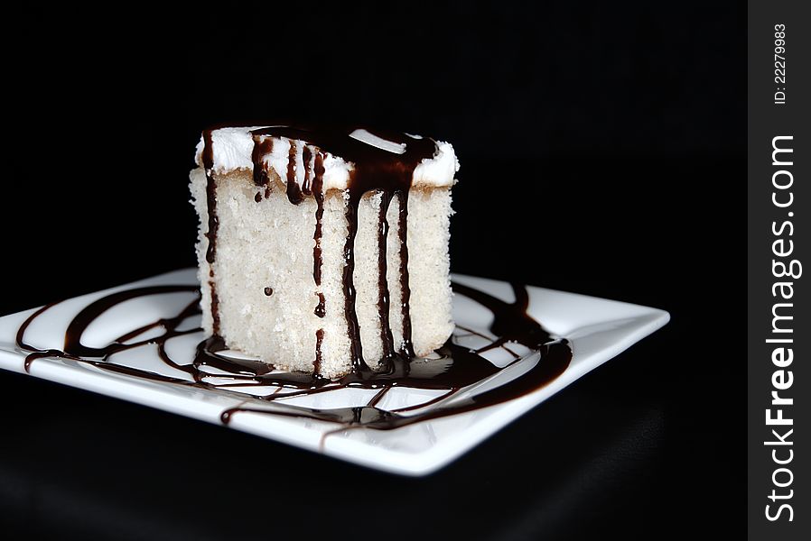 White cake with white icing that has been drizzled with chocolate fudge on a square white plate with a black background. White cake with white icing that has been drizzled with chocolate fudge on a square white plate with a black background.