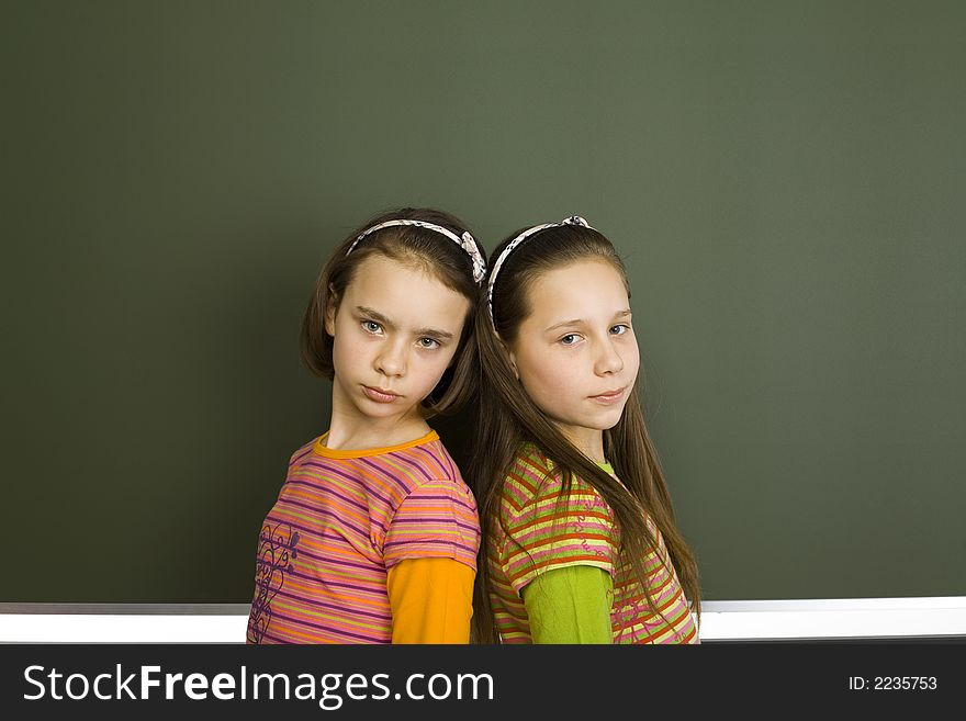 Two girls (11-13yo) are standing and looking at camera. There's blank greenboard behind them. Two girls (11-13yo) are standing and looking at camera. There's blank greenboard behind them.