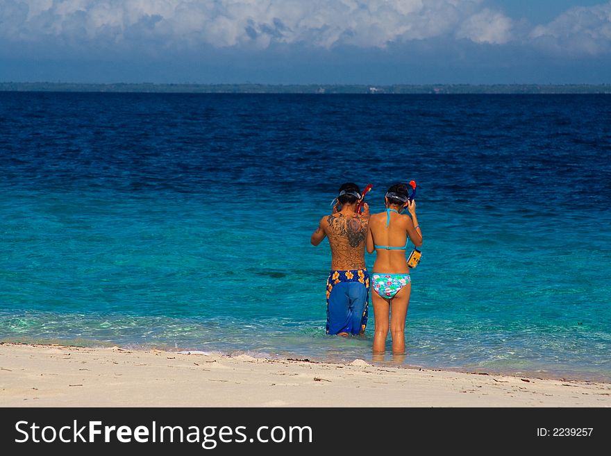 Two snorkelers prepare to swim in blue crystalline water beside a white tropical beach. Two snorkelers prepare to swim in blue crystalline water beside a white tropical beach