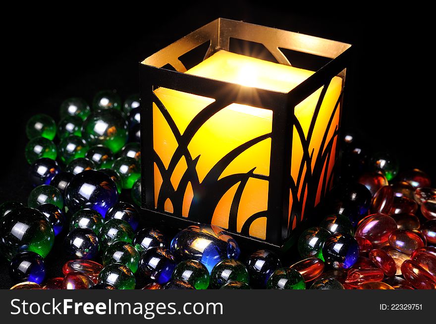 A yellow candle in a black metal candleholder burning in the dark with decorative multicolored glass stones. A yellow candle in a black metal candleholder burning in the dark with decorative multicolored glass stones