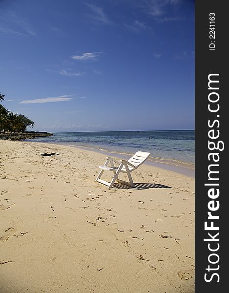 An empty white beach louge-style chair on a beach near the water against a blue sky background. An empty white beach louge-style chair on a beach near the water against a blue sky background.