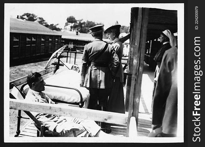 Queen Mary visiting a hospital on the Western Front. She is standing outside on a wooden balcony talking to a wounded soldier. He is lying on a bed with a sunshade propped up beside his head, to protect his face from the sun. There is a man sitting on a deckchair at the end of the bed, possibly another patient. A uniformed man stands next to the Queen, and a nurse watches the scene from the background. 

King George V and Queen Mary visited the Western Front on a number of occasions during World War I. Queen Mary spent a lot of her time visiting sick and wounded soldiers in hospitals behind the front line. An important function of royal visits was to raise the spirits of those involved in every aspect of the war.

[Original reads: .&#x27;.ON THE BRITISH WESTERN FRONT. THE VISIT OF HER MAJESTY THE QUEEN. - THE QUEEN CHATS WITH A PATIENT.&#x27;]

digital.nls.uk/74546706