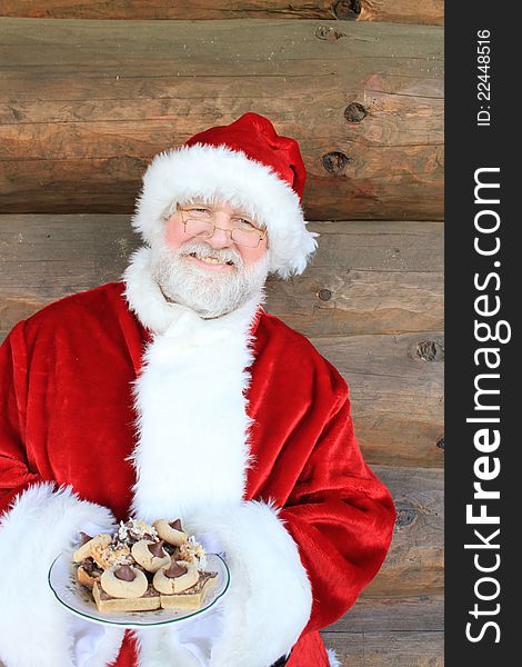 Santa standing in front of a log cabin wall holding a plate of Christmas cookies. Santa standing in front of a log cabin wall holding a plate of Christmas cookies.