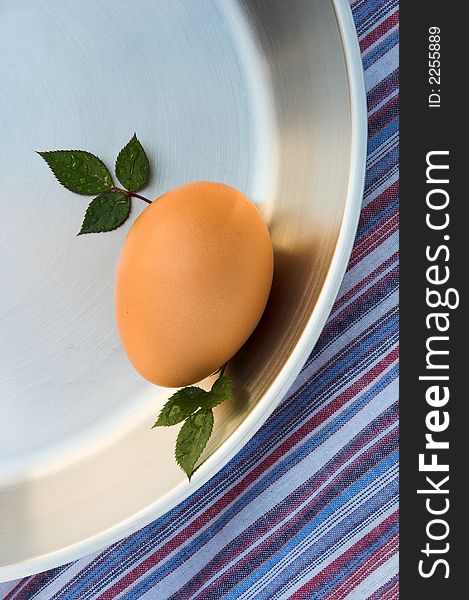 Chicken's egg in the shell in a silver frying pan. The image is shot from above. The pan is on a striped red, blue and white tablecloth. There are miniature rose leaves placed next to the egg. Chicken's egg in the shell in a silver frying pan. The image is shot from above. The pan is on a striped red, blue and white tablecloth. There are miniature rose leaves placed next to the egg