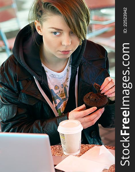 Portrait of blond stylish girl with cup and muffin on the terrace of the fast food with laptop on the table