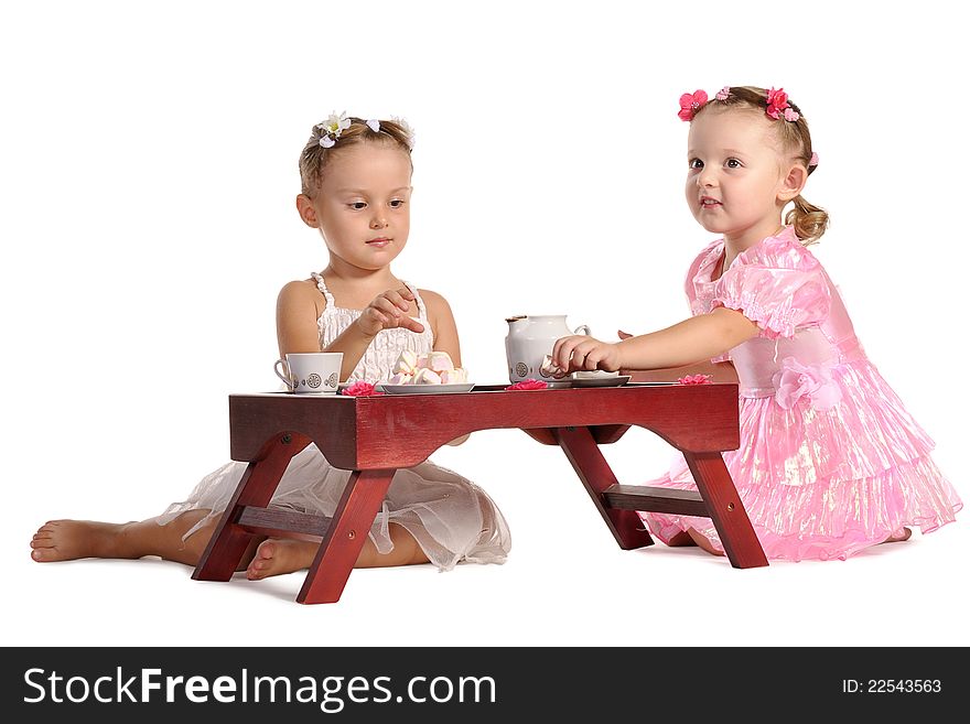 Two pretty twins sisters in beautiful dresses having tea wtih zephyr sitting at coffee table isolated on white background. Two pretty twins sisters in beautiful dresses having tea wtih zephyr sitting at coffee table isolated on white background