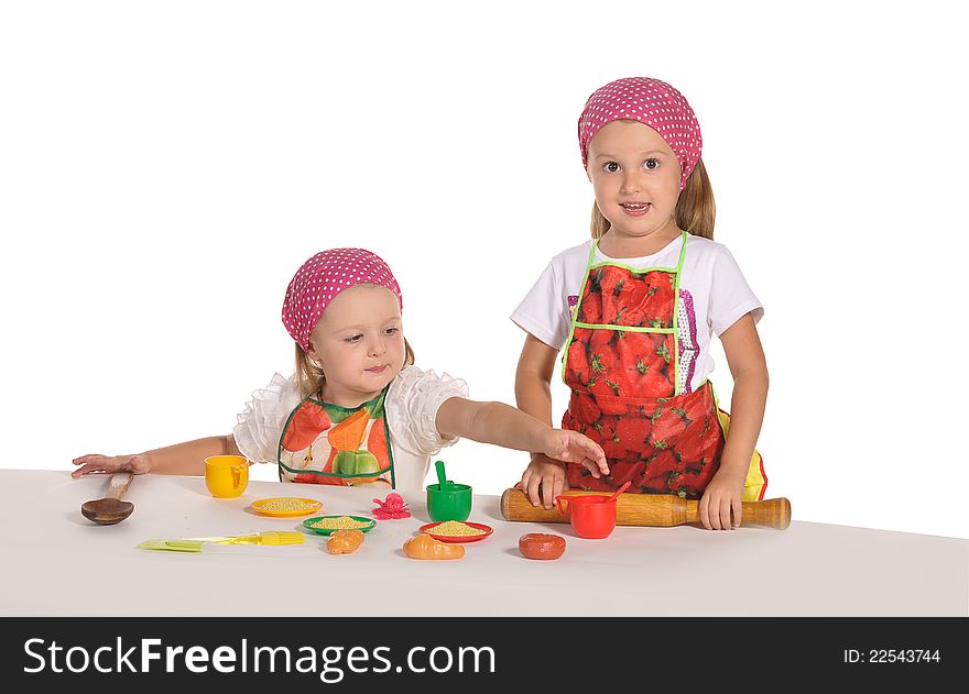 Two pretty little twins sisters wearing spotted pink headscarfs and colourful aprons pretending housewifes cooking isolated on white background. Two pretty little twins sisters wearing spotted pink headscarfs and colourful aprons pretending housewifes cooking isolated on white background