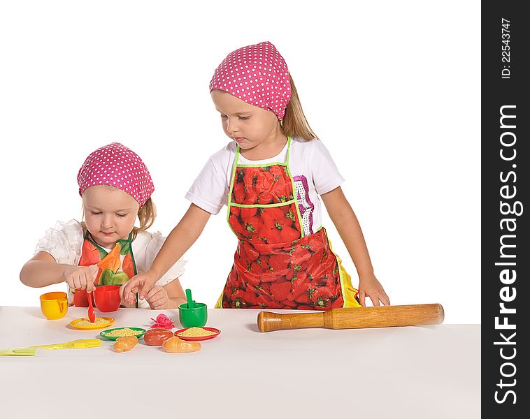 Two pretty little twins sisters wearing spotted pink headscarfs and colourful aprons pretending housewifes cooking isolated on white background. Two pretty little twins sisters wearing spotted pink headscarfs and colourful aprons pretending housewifes cooking isolated on white background