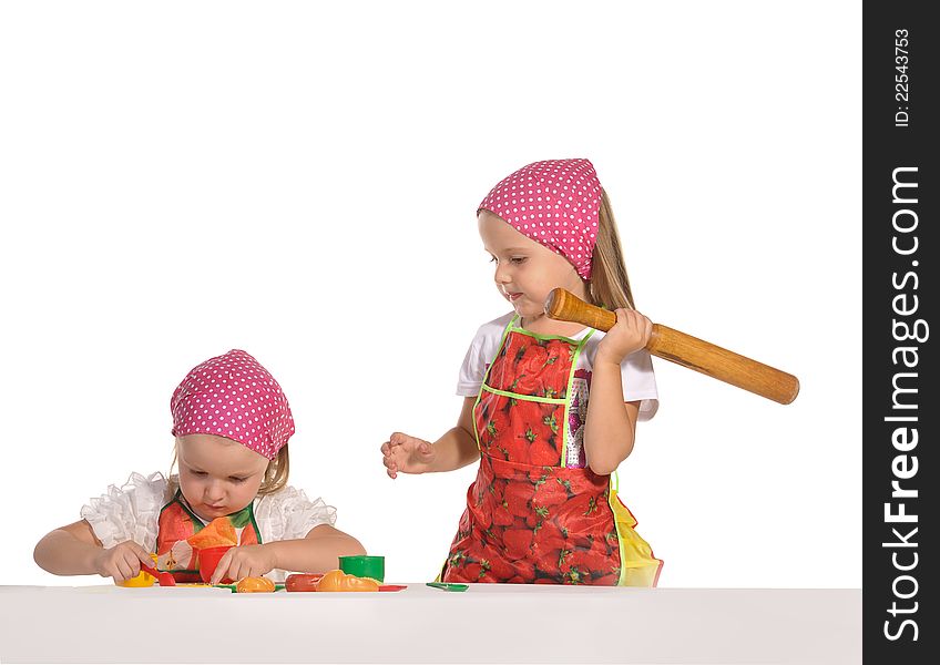 Two pretty little twins sisters wearing spotted pink headscarfs and colourful aprons pretending housewifes cooking isolated on white background. Two pretty little twins sisters wearing spotted pink headscarfs and colourful aprons pretending housewifes cooking isolated on white background