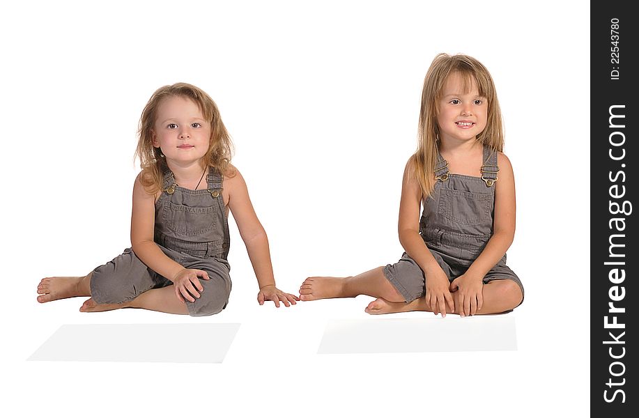 Two pretty twins sisters sitting on the floor barefooted and wearing rompers isolated on white background. Two pretty twins sisters sitting on the floor barefooted and wearing rompers isolated on white background