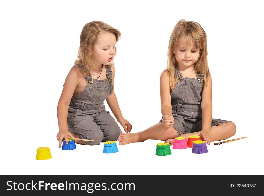 Two pretty barefooted twins sisters in rompers choosing paints isolated on white background. Two pretty barefooted twins sisters in rompers choosing paints isolated on white background
