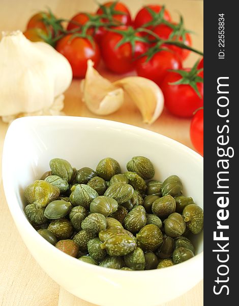 Pickled caper buds in a white bowl with tomatoes and garlic cloves at the background. Pickled caper buds in a white bowl with tomatoes and garlic cloves at the background