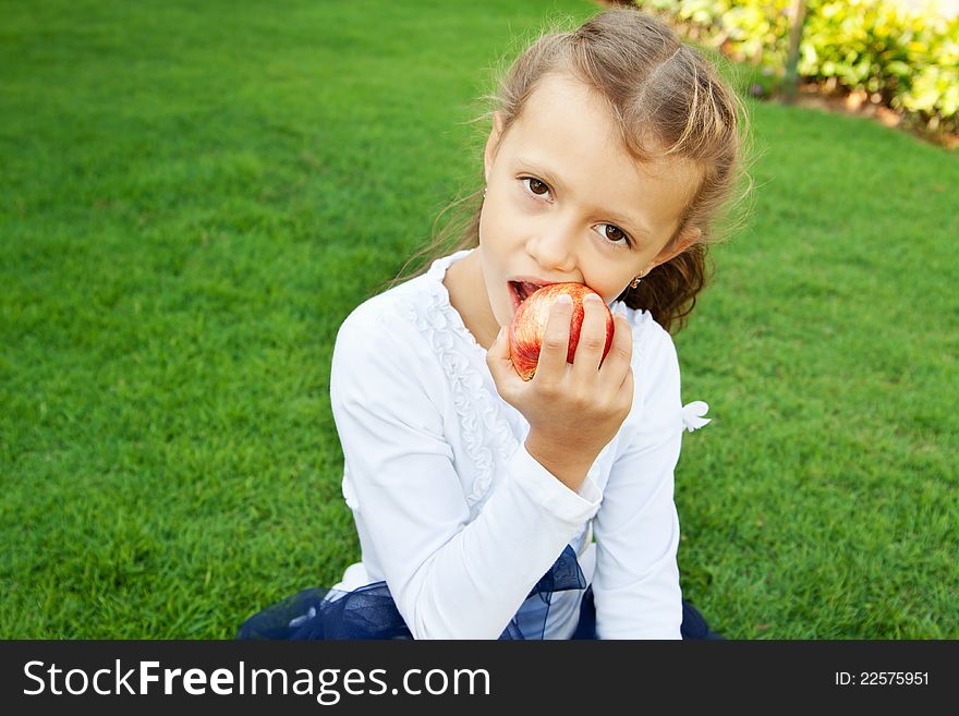 A pretty girl with long hair, eating red apple sitting on green grass. A pretty girl with long hair, eating red apple sitting on green grass