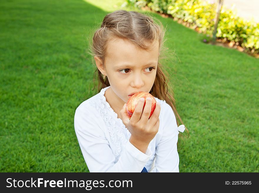 A pretty girl with long hair, eating red apple sitting on green grass. A pretty girl with long hair, eating red apple sitting on green grass
