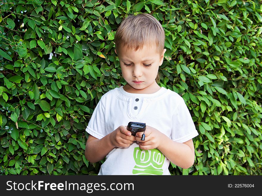 Cute boy playing with a mobile phone on the background of green leaves bush. Cute boy playing with a mobile phone on the background of green leaves bush