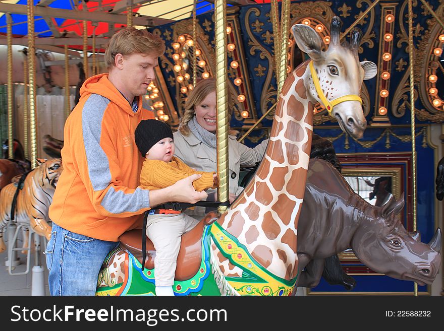 A dad and mom help their baby as he takes his first carousel ride on a giraffe. A dad and mom help their baby as he takes his first carousel ride on a giraffe.