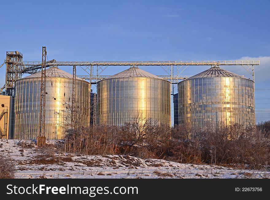 Group of silos filled with cereal grain against blue sky, placed in ploughed acres