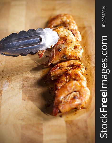 Tongs pick up grilled shrimp on wood plate. Tongs pick up grilled shrimp on wood plate.