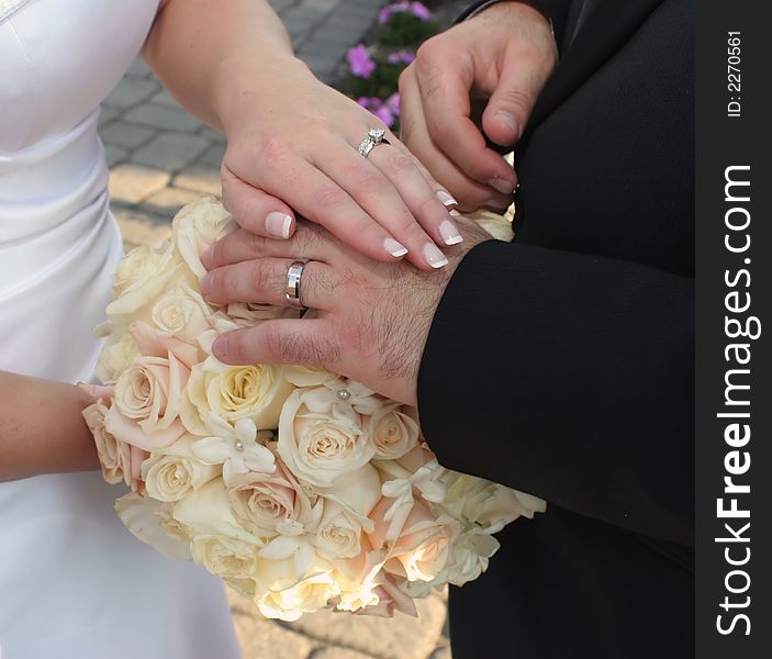 Detail of bride and groom's hands laid over bouquet of roses displaying wedding ring set. Detail of bride and groom's hands laid over bouquet of roses displaying wedding ring set