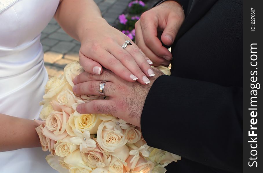 Detail of bride and groom's hands laid over bouquet of roses displaying wedding ring set. Detail of bride and groom's hands laid over bouquet of roses displaying wedding ring set