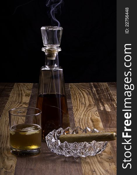 Smoking cigar in ashtray with decanter and glass on whiskey sitting on wooden table. Smoke curling around top of decanter. Dark background providing good contrast for wisps of smoke. Smoking cigar in ashtray with decanter and glass on whiskey sitting on wooden table. Smoke curling around top of decanter. Dark background providing good contrast for wisps of smoke.