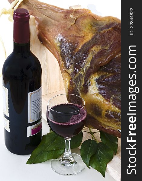 A front leg Serrano ham on displayed on a wooden rack and a bottle of red wine with a full glass of wine on a white background. A front leg Serrano ham on displayed on a wooden rack and a bottle of red wine with a full glass of wine on a white background.