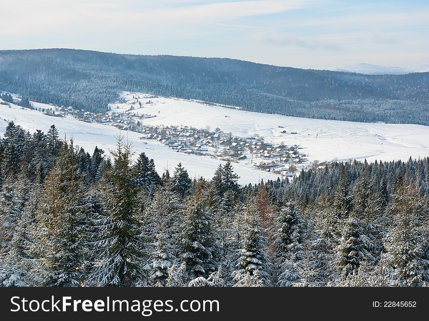 Snow village in valley between winter mountains with fir tree forest in foreground