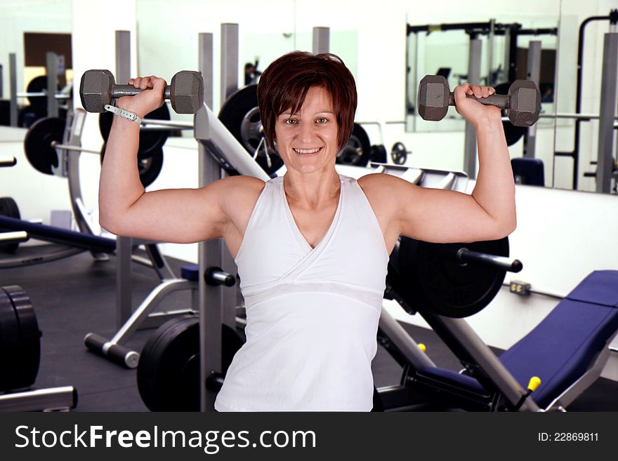 Woman in gym lifting dumbells over her head with gym in background. Woman in gym lifting dumbells over her head with gym in background.