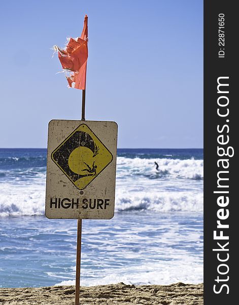 A High Surf sign on a beach warns of dangerous conditions as a surfer rides a wave in the background. A High Surf sign on a beach warns of dangerous conditions as a surfer rides a wave in the background.