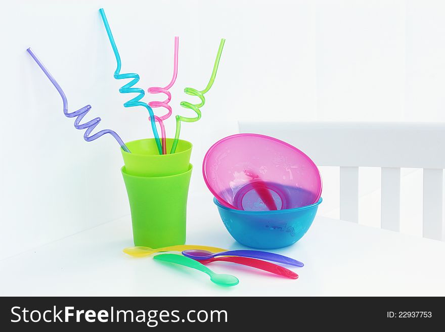 A high key image of brightly coloured kids cups, bowls, spoons and straws on a white table a chair. A high key image of brightly coloured kids cups, bowls, spoons and straws on a white table a chair.