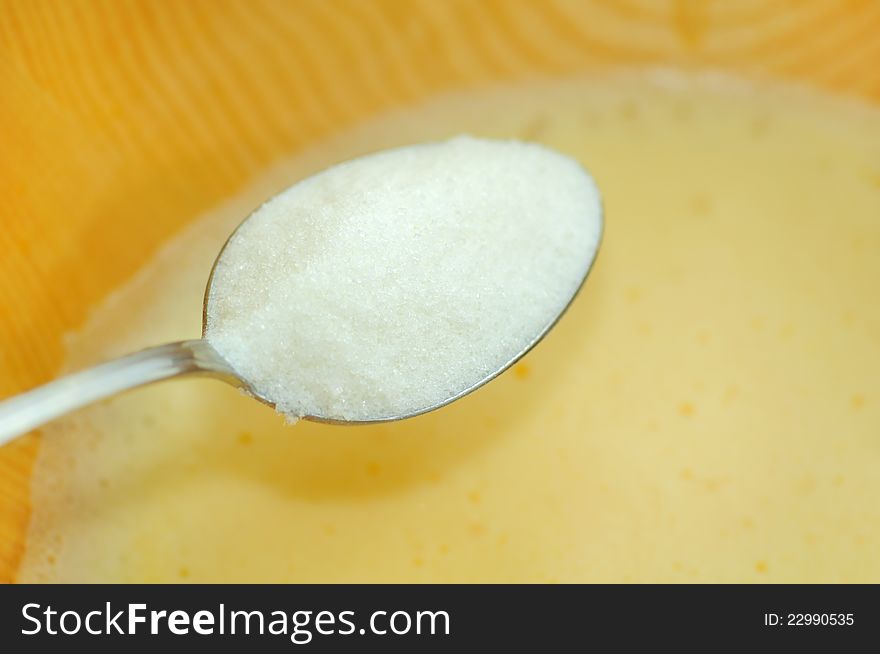 A table spoon of white sugar being added into a bowl with whipped eggs. A table spoon of white sugar being added into a bowl with whipped eggs
