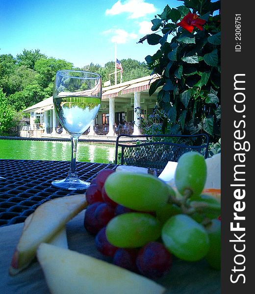 Fruit and cheese platter and glass of white wine arranged on table of restaurant terrace by a green lake with restaurant building in the background on a beautiful summer’s day. Fruit and cheese platter and glass of white wine arranged on table of restaurant terrace by a green lake with restaurant building in the background on a beautiful summer’s day